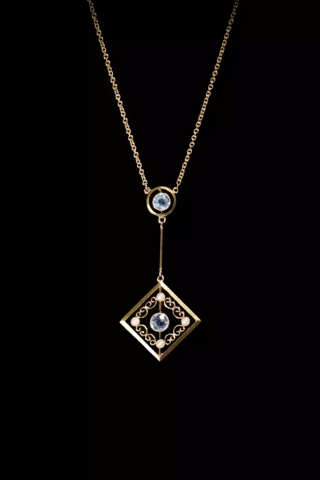 Gold necklace with a square pendant featuring intricate designs and blue gemstones, set against a black background.