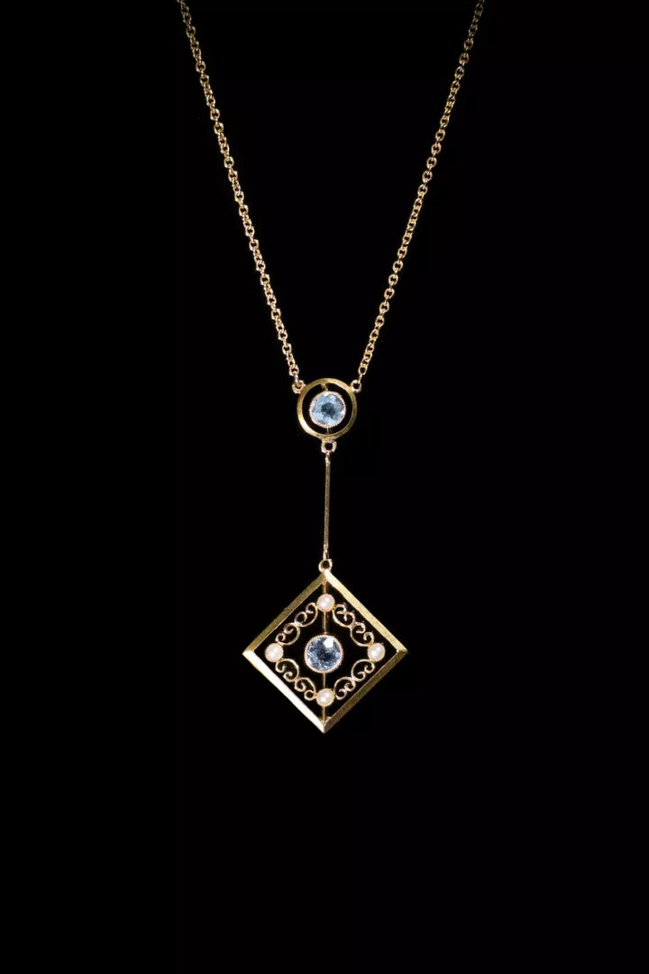 Gold necklace with a square pendant featuring intricate designs and blue gemstones, set against a black background.