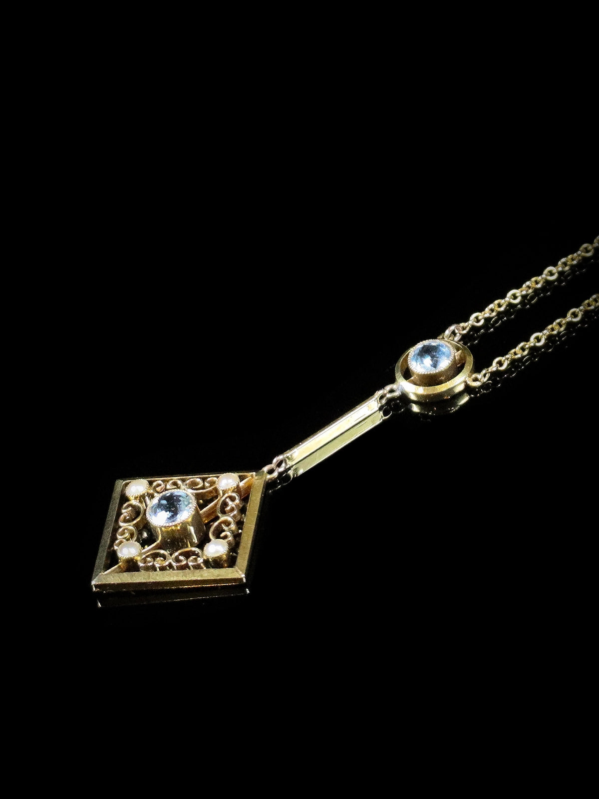 A gold necklace with a square, ornate pendant featuring a blue gemstone, suspended by a chain with an additional blue gemstone accent. The background is black.