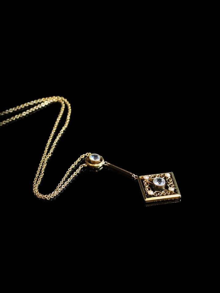 Gold necklace with a square pendant featuring a central gemstone, displayed on a black background.