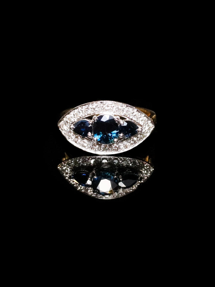 The 18ct Gold Three-Stone Sapphire Ring with a Diamond Halo is reflected on a black surface.