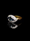 18ct gold sapphire ring with a large dark sapphire gemstone framed by a halo of small diamonds, reflecting on a black surface.