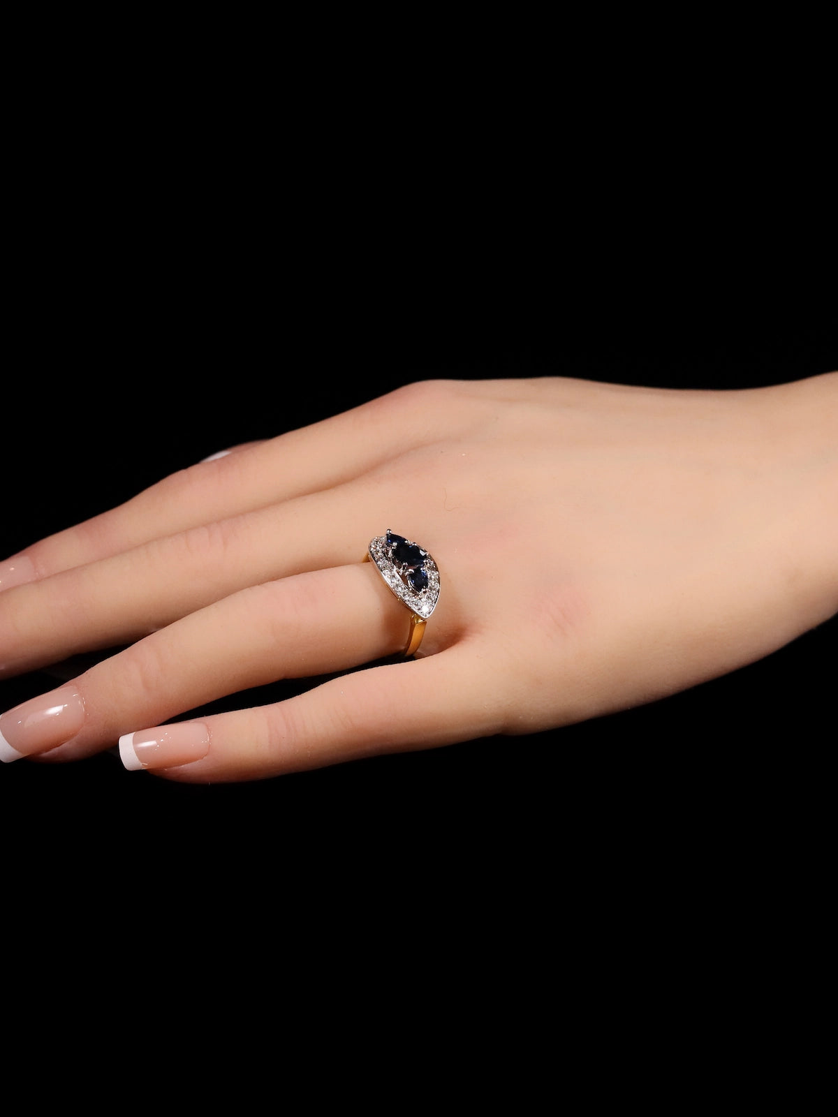 The image showcases a hand displaying the 18ct Gold Three-Stone Sapphire Ring with a dark central gemstone, featuring a diamond halo on a gold band, set against a black background.