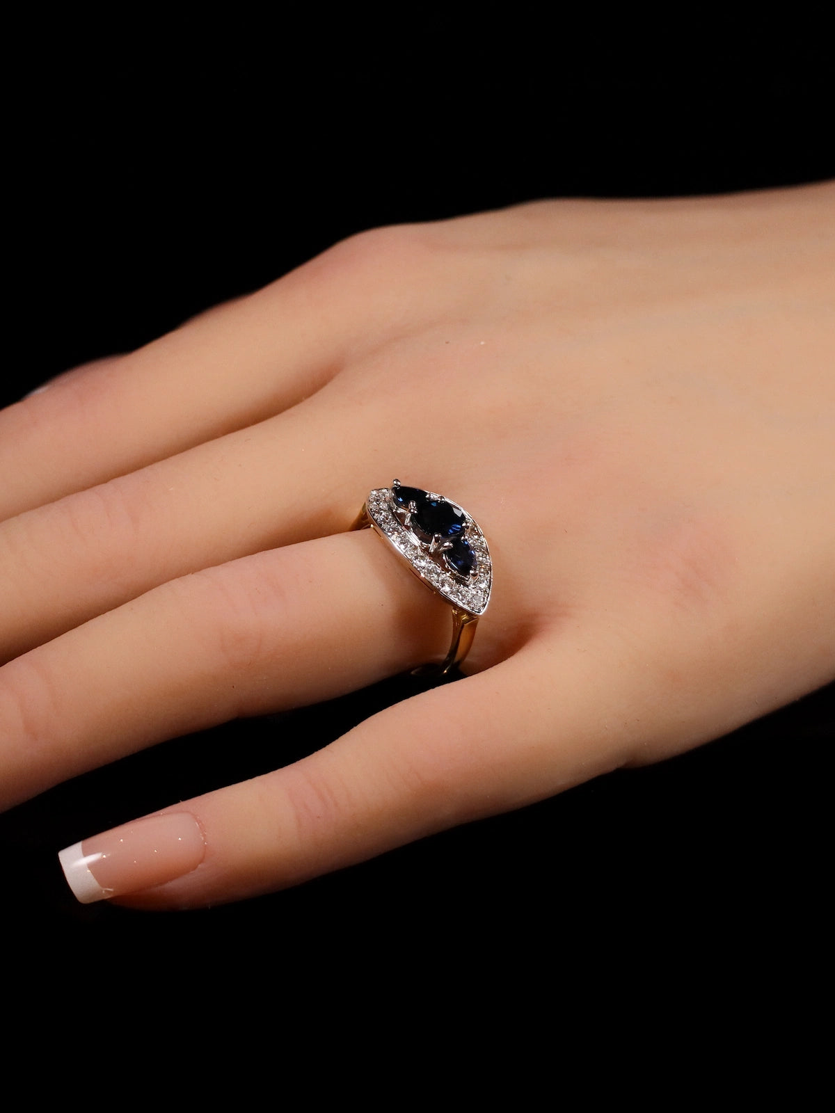A hand displays an 18ct gold marquise-shaped ring featuring a dark sapphire gemstone at the center, encircled by a halo of small clear diamonds.