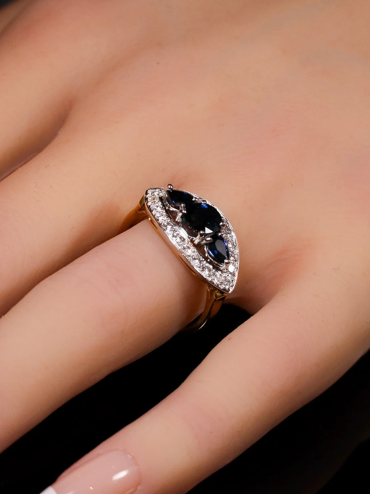 A hand wearing the 18ct Gold Three-Stone Sapphire Ring featuring a large, dark sapphire gemstone surrounded by a halo of smaller clear diamonds.