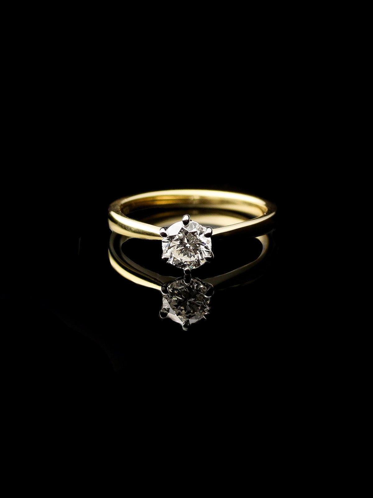 The 18ct Yellow Gold Round Brilliant 0.70ct Diamond Solitaire Ring is set against a black background, highlighting its reflective surface beneath.