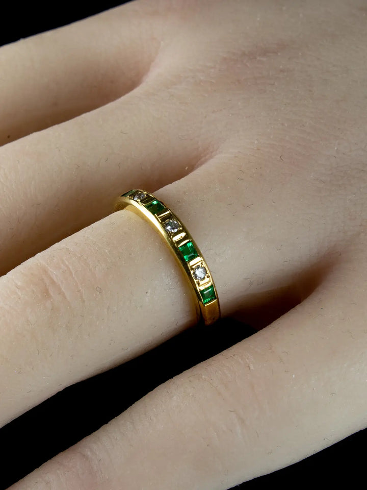 The Vintage 18ct Gold Emerald and Diamond Half Eternity Ring, featuring alternating green emeralds and clear diamonds, is worn on a finger.