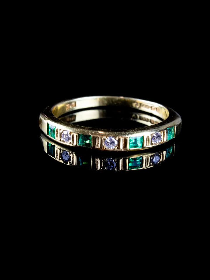Vintage 18ct gold ring featuring emerald and diamond gemstones set in a line, reflecting on a black surface.