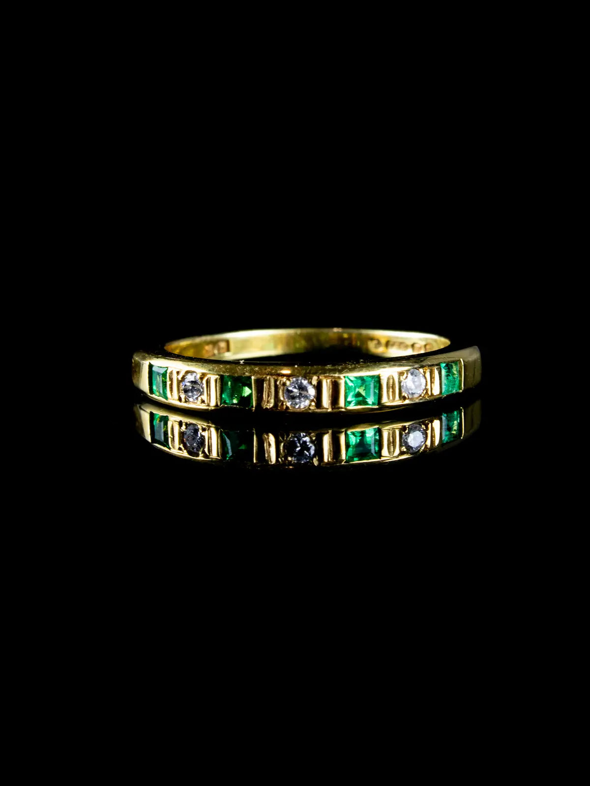 The Vintage 18ct Gold Emerald and Diamond Half Eternity Ring showcases a gold band adorned with alternating emerald and diamond stones, elegantly displayed on a reflective black surface.
