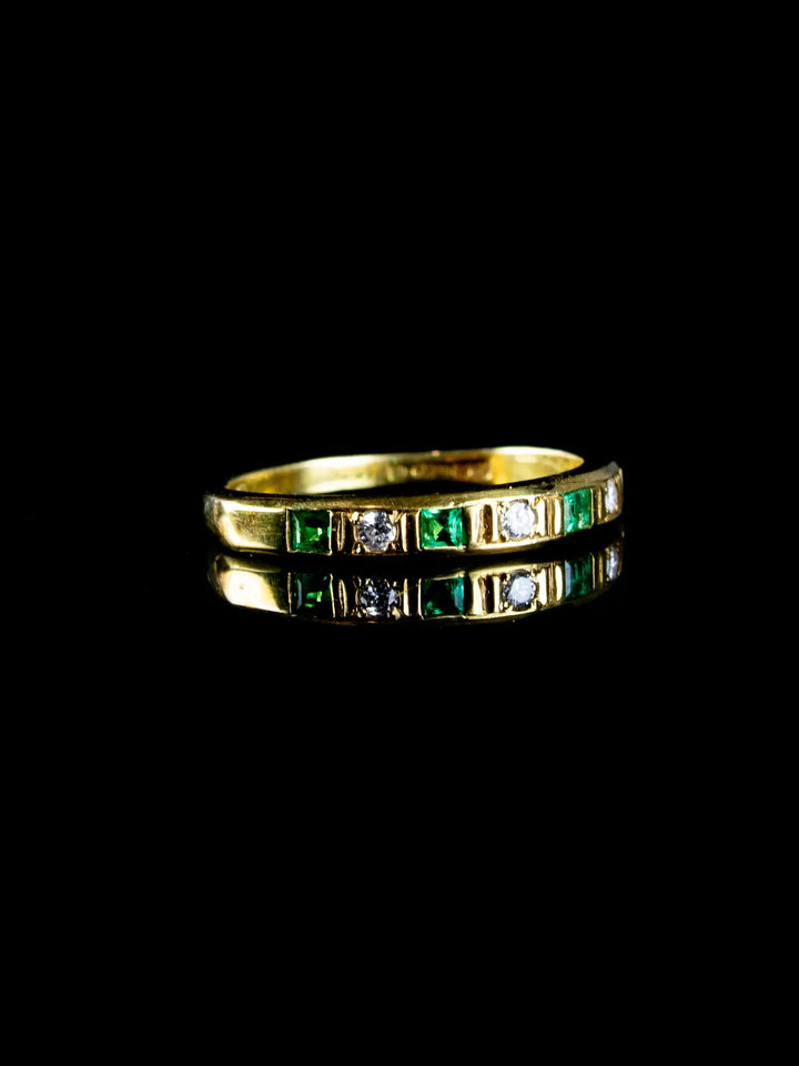 A Vintage 18ct Gold Emerald and Diamond Half Eternity Ring, featuring alternating green emeralds and clear diamonds, beautifully reflected on a black surface.