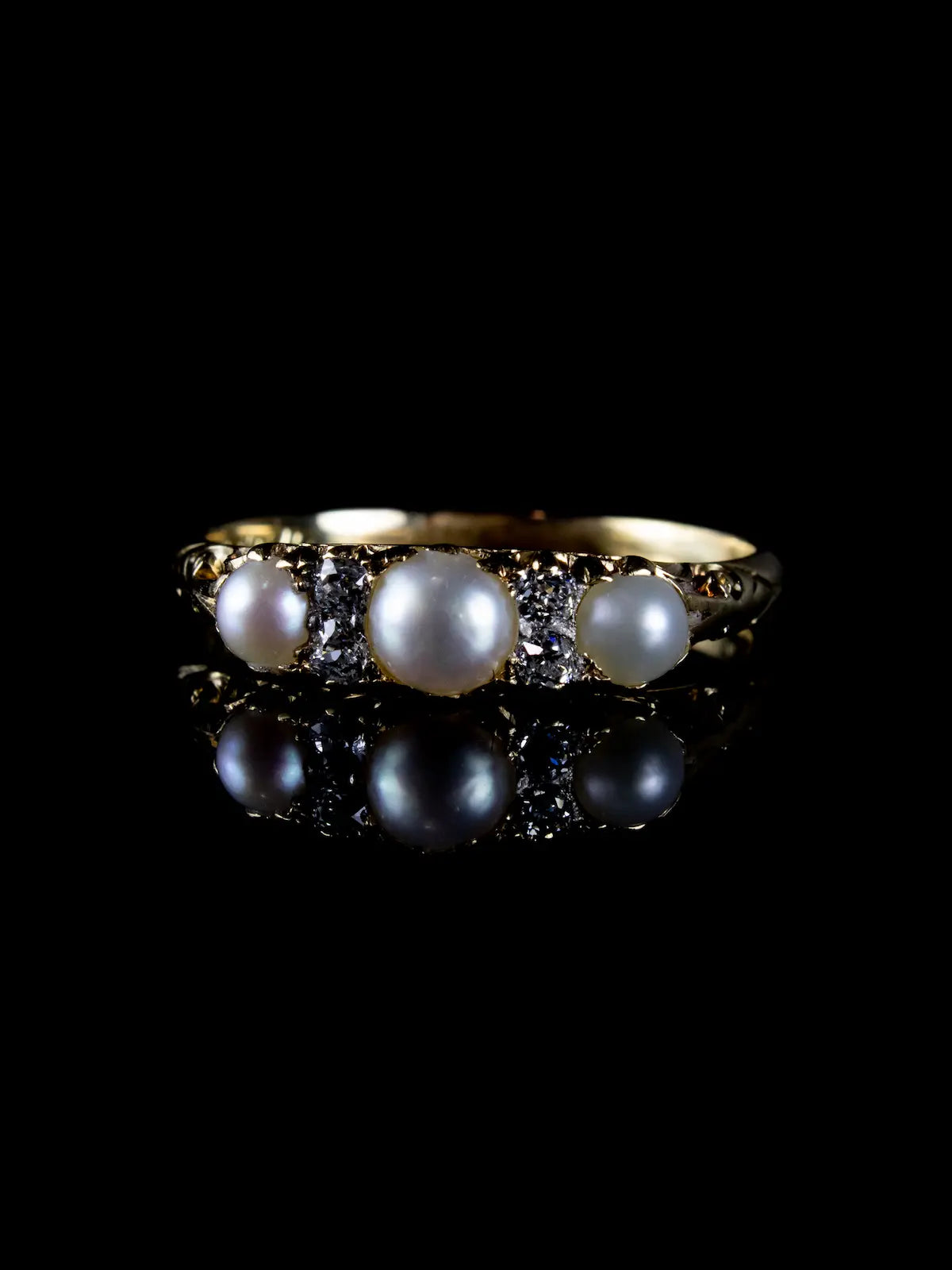 The Antique Victorian Seven Stone Pearl and Diamond Ring, Circa 1880, featuring a gold band with three round pearls and four small diamonds, is showcased against a black background with its reflection below.