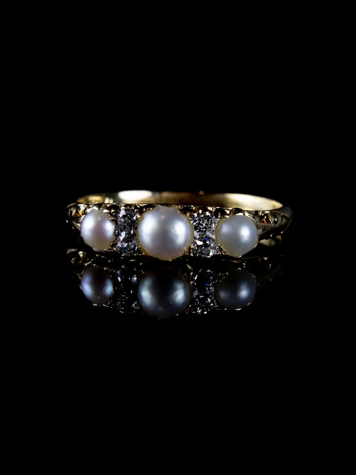 The Antique Victorian Seven Stone Pearl and Diamond Ring, Circa 1880, featuring a gold band with three round pearls and four small diamonds, is showcased against a black background with its reflection below.