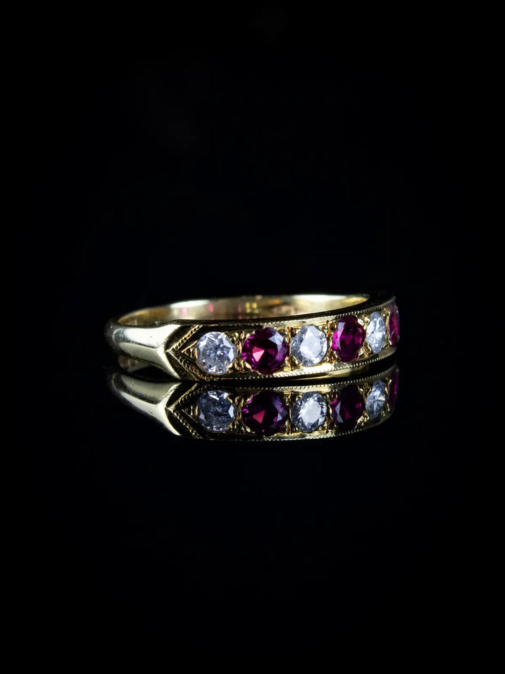 An 18ct gold seven stone ruby and diamond half eternity ring on a black reflective surface.