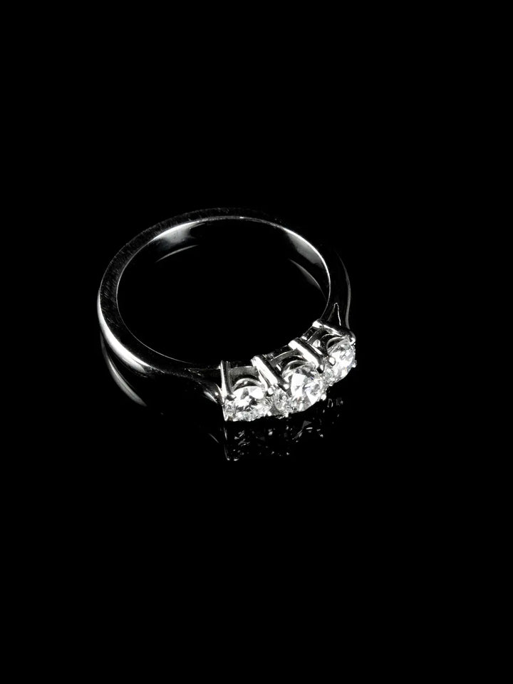 A platinum ring featuring three brilliantly clear diamonds set in a row, elegantly displayed on a black reflective surface.